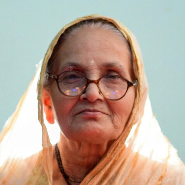 A senior woman in traditional clothes sits on a bed with a calm demeanor in a brightly lit room.